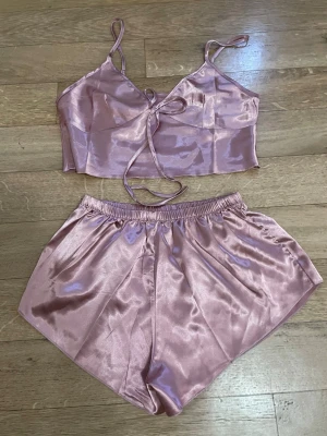 Rosa satinsätt med topp och shorts - Säljer ett glansigt rosa satinsätt med en croppad topp med smala axelband och knytning fram samt matchande shorts med resår i midjan. Perfekt för en chill kväll hemma eller som sovplagg. Materialet är mjukt och följsamt.