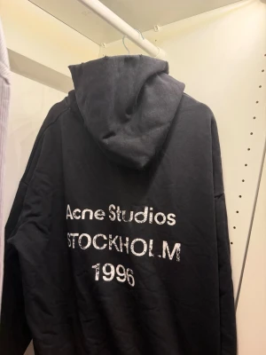 Svart hoodie från Acne Studios - Svart hoodie från Acne Studios med tryck på ryggen där det står 'Acne Studios STOCKHOLM 1996'. Klassisk passform med huva och mjukt material, perfekt för en chill och stilren look.