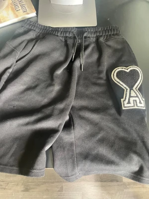 Ami Paris shorts  - Säljer ett par svarta shorts med snörning i midjan och en stor hjärtformad patch på ena benet. Shortsen har en avslappnad passform och är perfekta för chill dagar. Materialet känns mjukt och bekvämt, troligen bomull eller polyester.