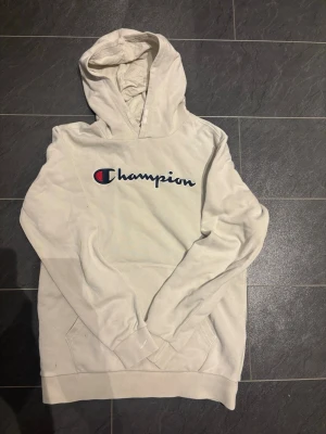 Vit hoodie från Champion  - Säljer en vit hoodie från Champion med stor broderad logga i blått, rött och vitt på bröstet. Hoodien har huva med dragsko och en klassisk magficka. Perfekt för en avslappnad och sportig stil. Hoodien behöver en tvätt!