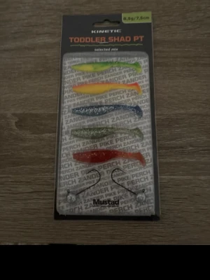 Kinetic Toddler Shad PT fiskedrag - Färgglada mjuka fiskedrag från Kinetic, modell Toddler Shad PT. Paketet innehåller flera drag i olika färger som grön, orange, blå, silver och röd samt två krokar från Mustad. Perfekt för gädda, abborre och gös.