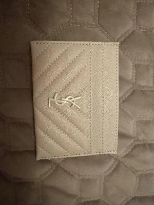 Beige kortfodral  - Snyggt beige kortfodral i strukturerat läder med beige YSL-logo på framsidan. Fodralet har ett stilrent, quiltat mönster och flera fack för kort. Perfekt accessoar för dig som gillar lyxig och minimalistisk design.