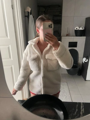 Vit teddyjacka med bröstfickor - Mysig vit teddyjacka i fluffigt material med två stora bröstfickor och krage. Jackan har dragkedja framtill och en avslappnad passform som ger en trendig look. Perfekt för att hålla värmen under kyliga dagar.