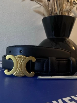 Svart bälte med guldspänne från Celine 25mm - 25mm och 90 cm långSnyggt svart bälte från Celine med ikoniskt guldfärgat spänne i form av deras Triomphe-logo. Bältet är gjort i slätt skinn och har klassisk design som passar till många olika outfits. Perfekt accessoar för att lyfta din stil. I går även en hålslagare 