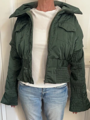 Grön cropped pufferjacka - Snygg grön cropped pufferjacka med hög krage och dragkedja framtill. Jackan har två bröstfickor med lock och coola quiltade detaljer på ärmar och midja. Perfekt för dig som vill ha en trendig och varm jacka med unik design.