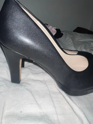 Svarta pumps från Clarks - Stilrena svarta pumps från Clarks med rundad tå och medelhög klack. Skorna är tillverkade i ett slätt material och har ljus insida med mjuk innersula för extra komfort. Perfekta för dig som vill ha en klassisk och tidlös look.