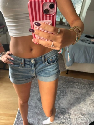 Blå denimshorts med uppvik - Säljer ett par klassiska blå jeansshorts med uppvikta benslut och femficksmodell. Shortsen har låg midja och stängs med knapp och dragkedja. Perfekta för varma dagar och enkla att matcha med olika toppar.