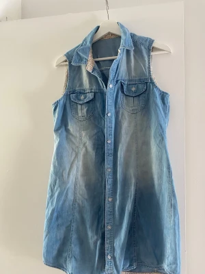 jeansklänning med knappar - Snygg  jeansklänning i ljusblå denim med knäppning framtill och två bröstfickor med lock. Klänningen har krage och en avslappnad passform, perfekt för en cool look. 