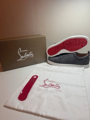 Gråa Christian Louboutin sneakers - Snygga gråa sneakers från Christian Louboutin med klassisk röd sula och vita kanter. Skorna har en låg profil, snörning framtill och är tillverkade i mocka. Perfekta för dig som vill ha något exklusivt och stilrent i garderoben.