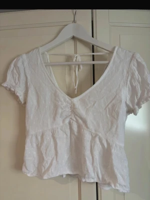 Vit blus med knyt från Hollister - Supersöt vit blus från Hollister med v-ringning, korta puffärmar och knyt i ryggen. Blusen har en lätt rynkad struktur och volang nedtill som ger en romantisk vibe. Perfekt till jeans eller kjol för en fräsch look. I storlek XS.