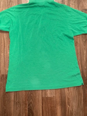 Grön pikétröja från Lacoste - Klassisk grön pikétröja från Lacoste med korta ärmar och den ikoniska krokodilloggan på bröstet. Tröjan har krage och knappar vid halsen. Perfekt för en avslappnad och stilren look.