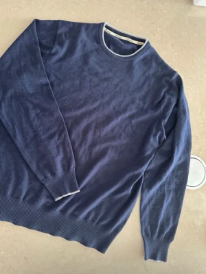 Mörkblå rundhalsad tröja - En stilren mörkblå tröja med rund halsringning och ribbade muddar vid ärmslut och nederkant.  Tröjan har en enkel design med kontrasterande kant vid halsen. Perfekt för dig som gillar klassiska och tidlösa plagg.ull