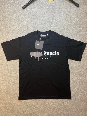 Svart Palm Angels t-shirt med tryck - Svart t-shirt från Palm Angels med klassiskt tryck framtill där det står 'Angels Paris' och en grå, drip-effekt över loggan. T-shirten har rund hals och korta ärmar. Perfekt för en streetwear-look.