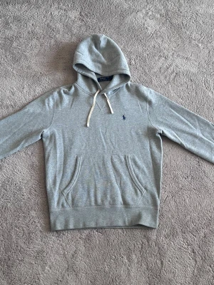Grå hoodie från Polo Ralph Lauren - Snygg grå hoodie från Polo Ralph Lauren med klassisk blå logga broderad på bröstet. Hoodien har huva med vita snören och en stor magficka. Perfekt för en chill och stilren look. Mjuk bomullsblandning som känns skön mot huden.