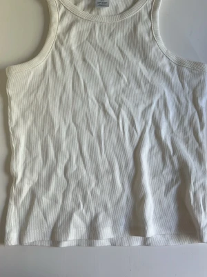 Vit ribbad linnetopp - Säljer en enkel och stilren vit linnetopp i ribbad bomull. Toppen är ärmlös med klassisk rund halsringning och har en tight passform. Perfekt att matcha med jeans eller kjol för en clean look.