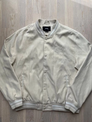 Pull & Bear collegejacka - beige, XL - Mycket bra skick. Inga tydliga tecken på användning Collegejacka från Pull & Bear, storlek XL, beige Full knappstängning, två fickor med knapp Beige- och vitrandiga muddar Bröstvidd: 61 cm Längd: 66 cm 100% polyester. Foder: 100% polyester Inga defekter