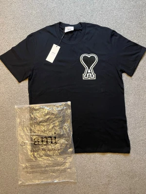 Ami Paris t shirt - Snygg svart t-shirt från Ami med stor vit och svart hjärtdetalj och bokstaven A på bröstet. Klassisk rund halsringning och korta ärmar. Tillverkad i mjuk bomull för skön känsla. Perfekt för dig som gillar stilrena plagg med en twist.