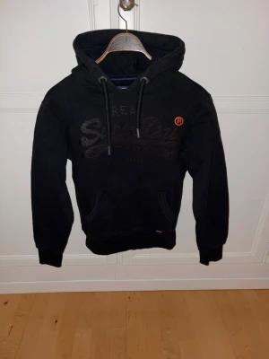 Svart hoodie Superdry - Säljer en svart hoodie från Superdry med stor logga på bröstet och en liten orange detalj. Hoodien har huva med dragsko, magficka och ribbade muddar vid ärmar och nederkant. Varm och skön hoodie