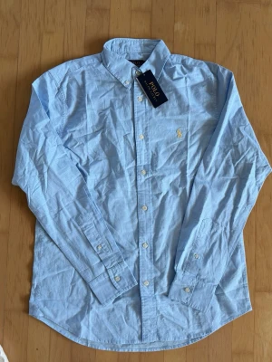 Ljusblå skjorta från Polo Ralph Lauren - Klassisk ljusblå skjorta från Polo Ralph Lauren med broderad logga på bröstet. Skjortan har lång ärm, knappar framtill och är tillverkad i bomull. Perfekt till jeans eller chinos för en clean och stilren look.