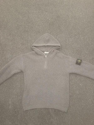 Grå stickad hoodie med dragkedja från Stone Island - Säljer en grå stickad hoodie med halv dragkedja framtill och ribbad struktur. Tröjan har en huva och en svart patch med logga på vänster ärm. Perfekt för dig som gillar streetwear och vill ha något både snyggt och bekvämt. Från stone island. ✅