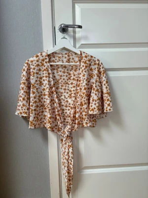 Två omlottoppar med knyt och mönster - Säljer två snygga omlottoppar med knyt framtill. Ena toppen är vit med brunt blommönster och den andra har ett patchworkmönster i brunt, orange och beige. Båda har vida ärmar och croppad passform, perfekta för sommarens alla outfits.