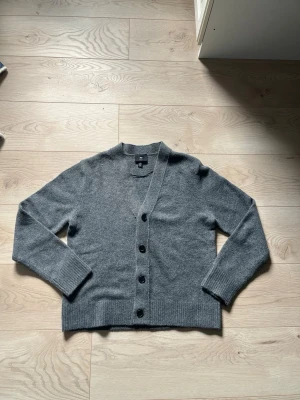 Grå kofta från H&M - Mysig grå kofta från H&M med rak passform och fyra knappar framtill. Koftan har långa ärmar, ribbade muddar och är tillverkad i ett mjukt material som passar perfekt till lager på lager. Enkel och stilren design som funkar till det mesta.