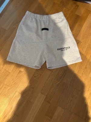 Grå shorts från Essentials Fear of God - Snygga ljusgrå shorts från Essentials Fear of God med elastisk midja och svart logga framtill. Perfekta för sommaren och har en clean, enkel design som passar till allt. Tillverkade i mjuk bomull för maximal komfort.