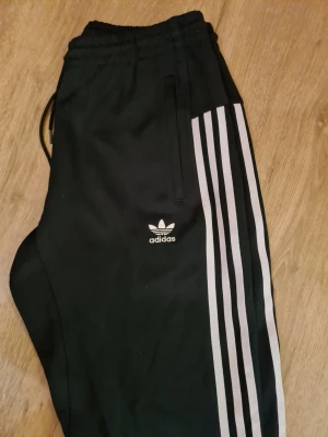 Svarta Adidas mjukisbyxor med ränder - Klassiska svarta mjukisbyxor från Adidas med de ikoniska tre vita ränderna längs benen och Adidas-logga på låret. Byxorna har resår i midjan med dragsko och sidofickor. Perfekta för chill eller träning. Korta.
