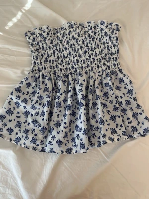 Blommig blåvit ärmlös topp - Supersöt ärmlös topp med blått blommönster på vit botten. Smockad upptill för en bekväm passform och volangkant upptill. Perfekt för varma dagar och ger en romantisk vibe till din outfit. Kan även användas som kjol.