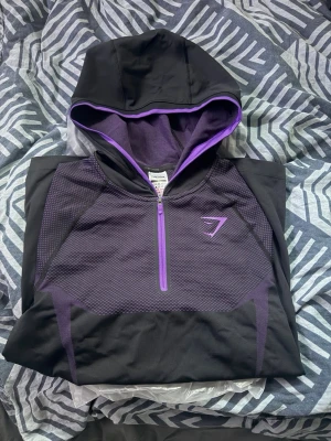 Gymshark Onyx V5 Ziphoodie Purple - Snygg svart hoodie från Gymshark med lila detaljer och halv dragkedja framtill. Hoodien har en sportig design med meshpaneler och en stor huva med lila kant. Perfekt för dig som gillar en aktiv stil och vill sticka ut.