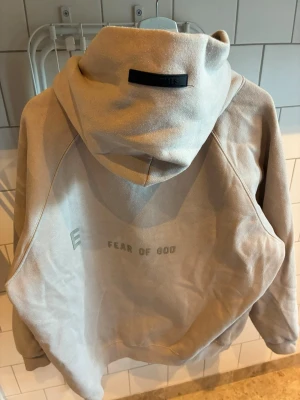 Fear of God ESSENTIALS - hoodie - Säljer en beige hoodie från Fear of God Essentials med tryck på ryggen. Hoodien har stor huva, känguruficka framtill och mjukt material som känns riktigt skönt. Perfekt för dig som gillar streetwear och vill ha en clean look.