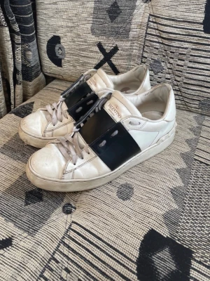 Valentino vita sneakers med svart band - Säljer ett par vita Valentino sneakers med ett brett svart band över vristen. Skorna har klassisk låg profil, vita snören och diskret logga på plösen. Tillverkade i läder med gummisula. Perfekta för dig som gillar stilrena och exklusiva sneakers.
