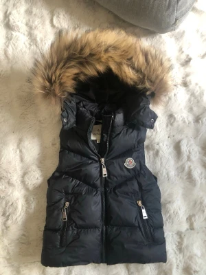 Barn svar  dunväst med äkta päls Moncler - Snygg svart dunväst från Moncler med avtagbar huva och stor päls runt kanten. Västen har två dragkedjefickor fram och Moncler-logga på bröstet. Perfekt för lager på lager och håller dig varm under kyliga dagar.
