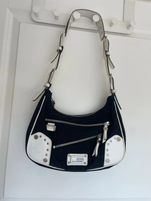 Svart och vit axelväska med detaljer - Cool svart axelväska med vita detaljer och silverfärgade nitar. Väskan har två snedställda dragkedjefickor framtill även en baktill, justerbart axelband och vita kanter. I väskan finns även tre fack. Materialet är en mix av textil och konstläder. Perfekt för dig som gillar en edgy och unik look. Väskan är använd och har några defekter (kolla bilderna) men de syns ej när du använder den! Köpt secondhand, Bara att fråga för mer info!