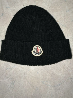 Svart Moncler mössa i ull - Snygg svart ribbstickad mössa från Moncler med klassisk uppvikt kant och broderad Moncler-logga framtill. Tillverkad i mjuk ull som håller dig varm och stilren under kalla dagar. Säljer den för använde aldrig mössa stylar håret istället så den har bara luggit i garderoben sen jag köpte den priset kan diskuteras👍