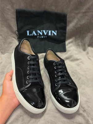 Svarta sneakers från Lanvin - Säljer ett par svarta sneakers från Lanvin med snygg kontrast mellan mocka och lackdetaljer på tån. Skorna har vita sulor och klassisk snörning. Perfekta för dig som vill ha en stilren och lyxig look.