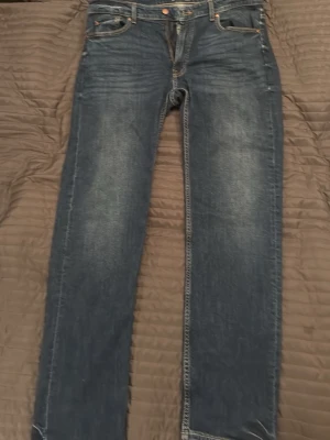 Blå raka jeans med slitningar - Säljer ett par klassiska blå jeans med raka ben och diskreta slitningar framtill. Jeansen har fem fickor, knappgylf och kontrastsömmar. Perfekta till en avslappnad stil och tillverkade i denim.