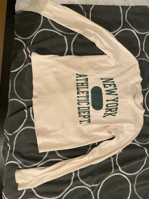 Vit långärmad tröja med tryck - Vit långärmad tröja med grönt tryck där det står 'NEW YORK ATHLETIC DEPT.' i stora bokstäver. Tröjan har rund halsringning och en avslappnad passform, perfekt för en sportig och casual stil.