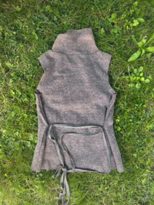 Zara topp -  brungrå stickad topp med hög krage och knytband i midjan. Västen är ärmlös och har en rak passform som kan justeras med knytet. Endast använd 1 gång