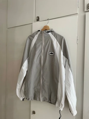 Cortiez windbreaker  - Snygg grå och vit vindjacka med dragkedja framtill och luva. Jackan har ett svartvitt märke på bröstet och justerbar nederkant med snörning. Perfekt för blåsiga dagar och enkel att matcha med olika outfits.