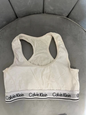 Vit sport-bh från Calvin Klein - Vit sport-bh från Calvin Klein med bred resår och logga runt om. Bh:n har brottarrygg och är tillverkad i mjukt, stretchigt material som ger en bekväm passform. Perfekt för en avslappnad och sportig stil.