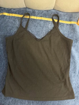 Svart ribbad linnetopp med spets - Snygg svart ribbad linnetopp med tunna axelband och v-ringning. Toppen har fina spetsdetaljer längs urringningen som ger en extra touch. Perfekt att styla till jeans eller kjol för en trendig look.