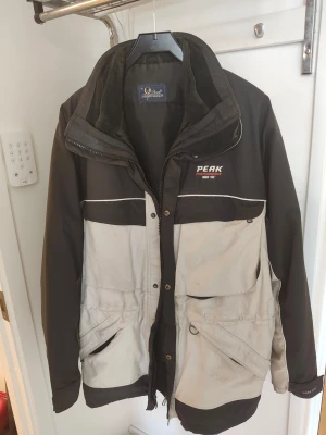 Svart Beige vindjacka från Peak Performance - Snygg svart vindjacka från Peak Performance med ljus insida och dragkedja. Jackan har diskret logga på bröstet och är tillverkad i ett lätt, vindtätt material. Perfekt för blåsiga dagar och enkel att matcha med olika outfits. Finns även avtagbar jacka på insidan. Jackan är Beige/Svart
