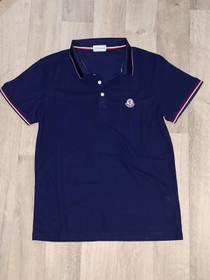 Mörkblå pikétröja från Moncler - Snygg mörkblå pikétröja från Moncler med korta ärmar och klassisk krage. Tröjan har vita och röda detaljer på krage och ärmslut samt Moncler-logga broderad på bröstet. Perfekt till jeans eller shorts för en sportig look. Passae också som S