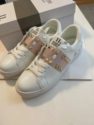 Vita sneakers med nitar och rosa band - Snygga vita sneakers med breda rosa band över vristen, dekorerade med guldiga fyrkantiga nitar. Skorna har vita snören och en chunky sula. Materialet ser ut att vara slätt läder eller konstläder. På plösen står det '11. Rockstud Untitled'.