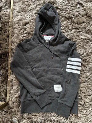  Thom Browne hoodie - Snygg grå hoodie från Thom Browne med klassiska vita ränder på ena ärmen och en patch med logga framtill. Hoodien har använts en gång och inget mer. Hör av er vid minsta lilla fundering!
