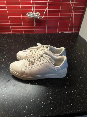 Vita sneakers från Tommy Hilfiger - Stilrena vita sneakers från Tommy Hilfiger med klassisk låg profil och snörning. Skorna är tillverkade i slätt lädermaterial och har diskret logga på sidan. Perfekta för en clean och enkel look.