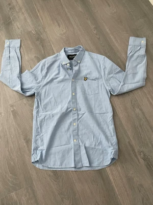 Ljusblå skjorta från Lyle & Scott - Klassisk ljusblå skjorta från Lyle & Scott med button-down krage och gul logga på bröstfickan. Skjortan har långa ärmar, vita knappar och är tillverkad i mjuk bomull. Perfekt för en clean och stilren look.