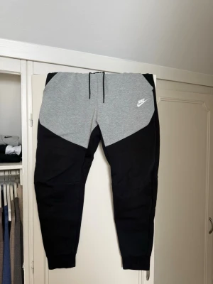 Nike mjukisbyxor svart/grå - Snygga mjukisbyxor från Nike i svart och grått med dragsko i midjan och ribbade muddar vid bensluten. Byxorna har en modern, avsmalnande passform och är tillverkade i mjukt sweatshirtmaterial. Nike-logga på vänster lår.