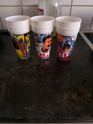 Plastmuggar med sportmotiv - Säljer ett set med sex vita plastmuggar med färgglada tryck av olika sportmotiv och tecknade figurer. Muggarna har olika illustrationer runt om och är perfekta för dryck vid filmkvällar eller temafester.